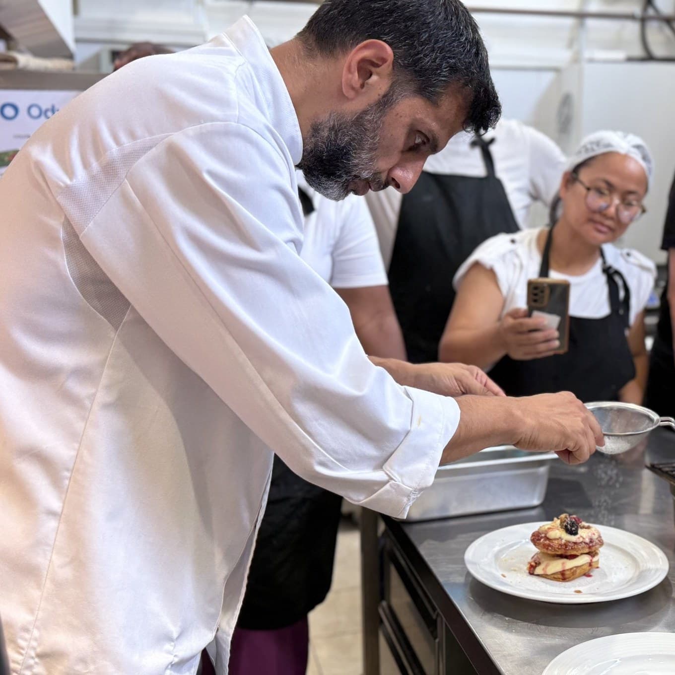 Ο επικεφαλής σεφ της GoodBite σερβίρει ένα επιδόρπιο στην κουζίνα της Odyssea Academy, ενώ εκπαιδευόμενοι παρακολουθούν